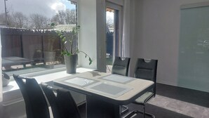 A modern room with a large window, a dining table with chairs, a potted plant, and a sliding door. - Comfort vacation apartment Niemann 4 with private garden/terrace (Lübeck)