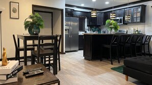 A modern kitchen with dark cabinetry, stainless steel appliances, and pendant lighting. There is a dining area with a table and chairs, and a living room with a sofa and a coffee table. - Secluded Contemporary 2BR 2BA Suite (Dundas Town)
