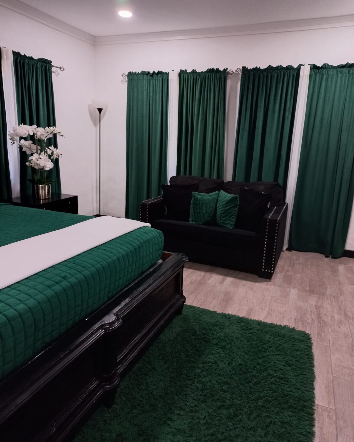 Quarto com cama, sofá, cortinas verdes e uma pequena mesa com flores.