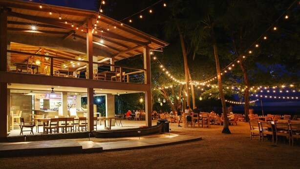 An outdoor dining area with string lights, wooden tables, and chairs, surrounded by palm trees. - Your Perfect Tropical Vacation Villa (Guatemala de Santa Rosa)