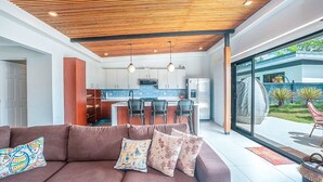 A modern living room with a brown sofa, a kitchen in the background, and a large sliding glass door leading to a garden. - Your Perfect Tropical Vacation Villa (Guatemala de Santa Rosa)