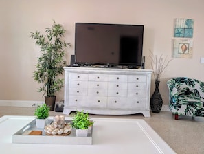A living room with a television on a white cabinet, a potted plant, a coffee table with decorative items, and a patterned armchair. - Dancing Seahorse Beach House with Heated Pool! (Largo)