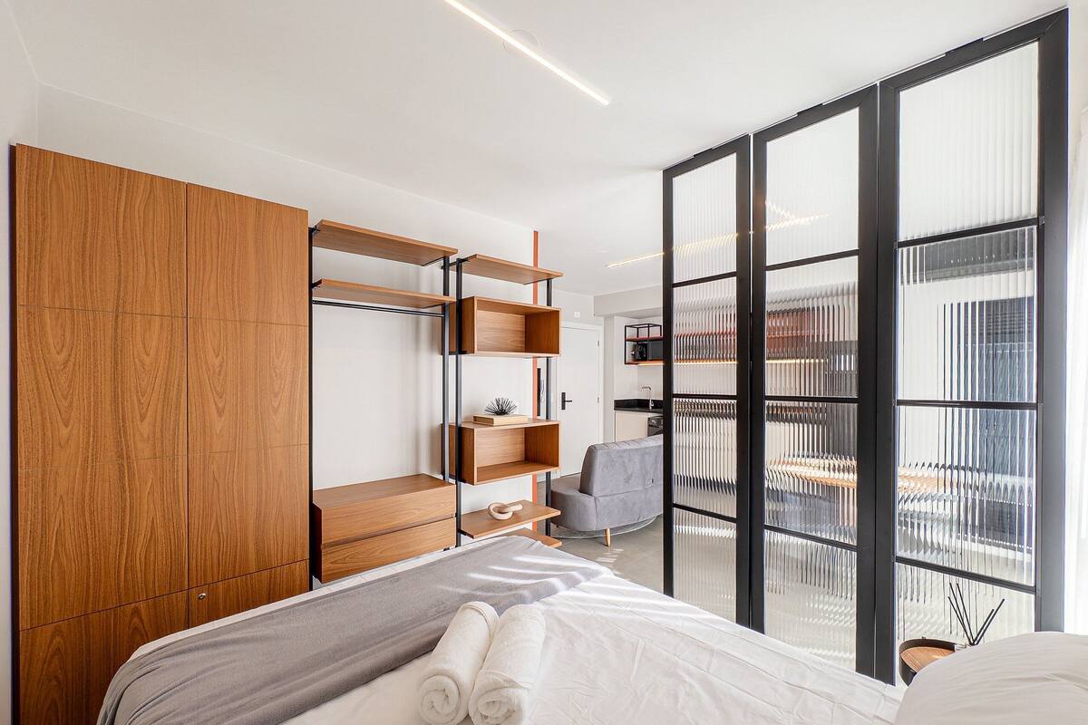 A modern hotel room with a bed, a wooden wardrobe, a grey sofa, and a balcony with a view.
