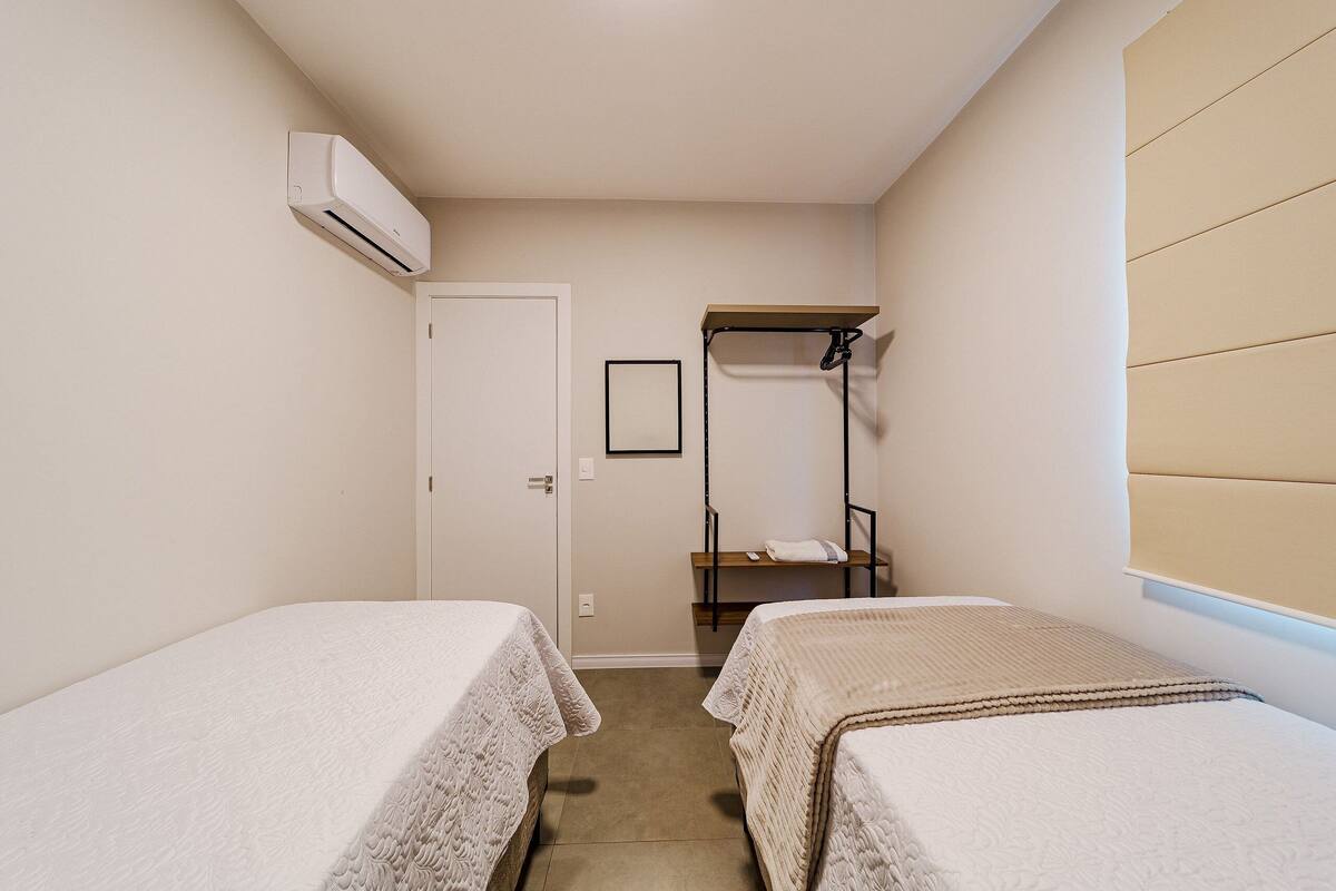 A small, clean room with two single beds, a door, a shelf, and an air conditioning unit.