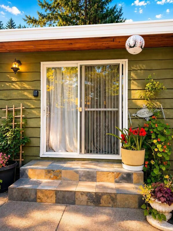Una casa con fachada verde, puerta corredera de vidrio, letrero de bienvenida y plantas en macetas.