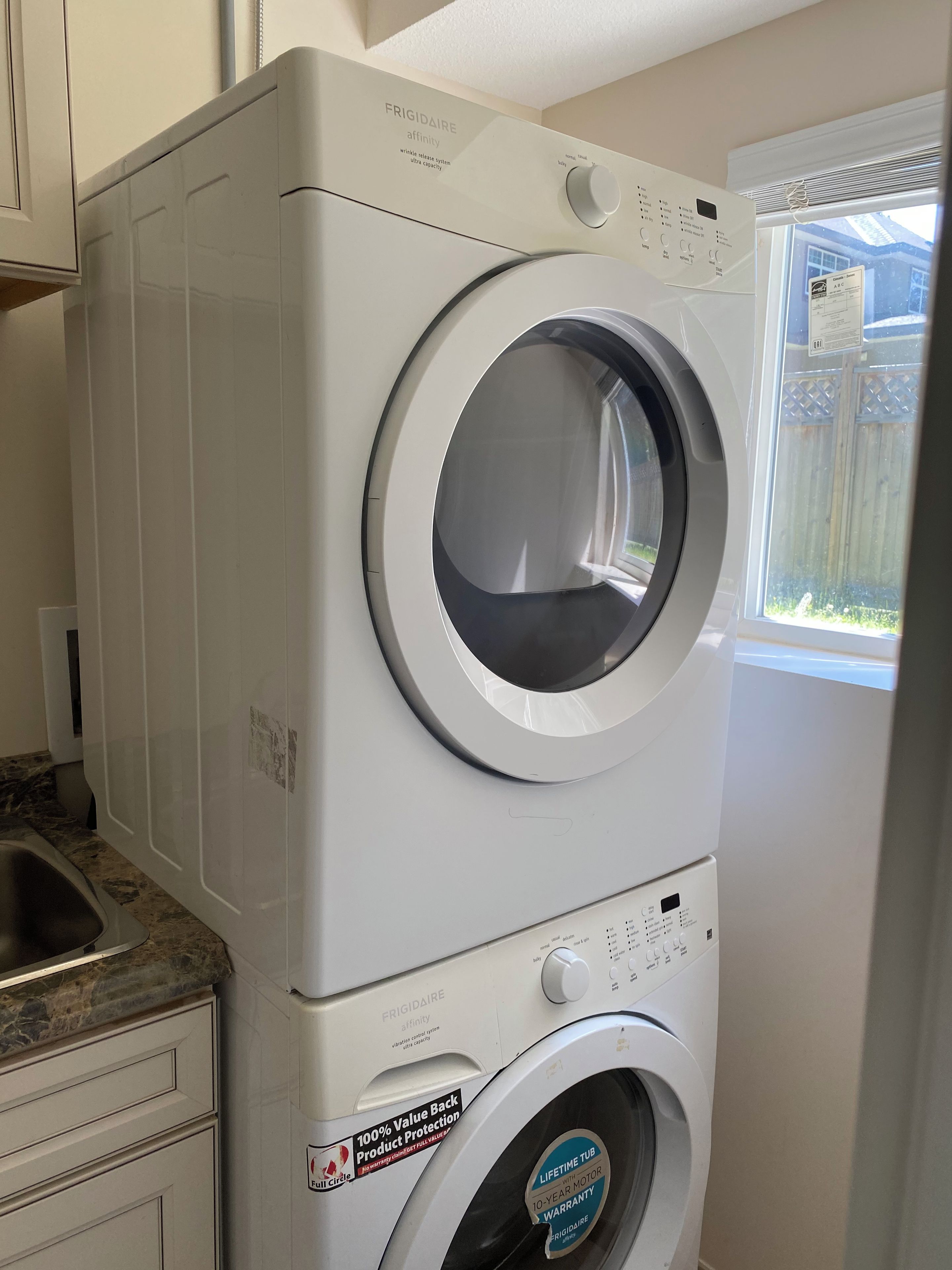 A front-loading Frigidaire washer and dryer unit with a stainless steel control panel, a 10-year motor warranty, and a 100% value back product protection sticker.