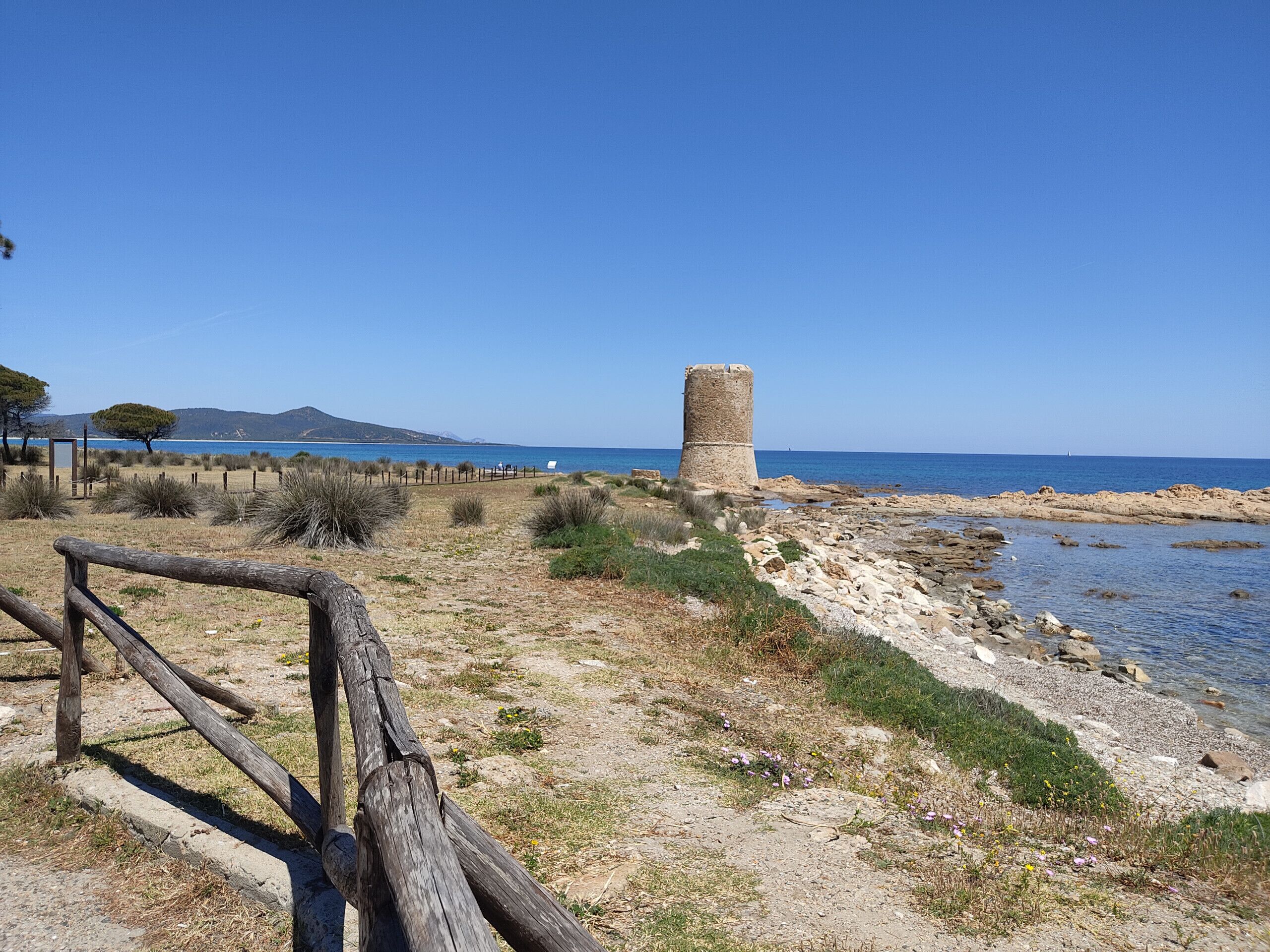 Un paisaje costero con una torre de piedra, playa arenosa y cielo azul despejado.