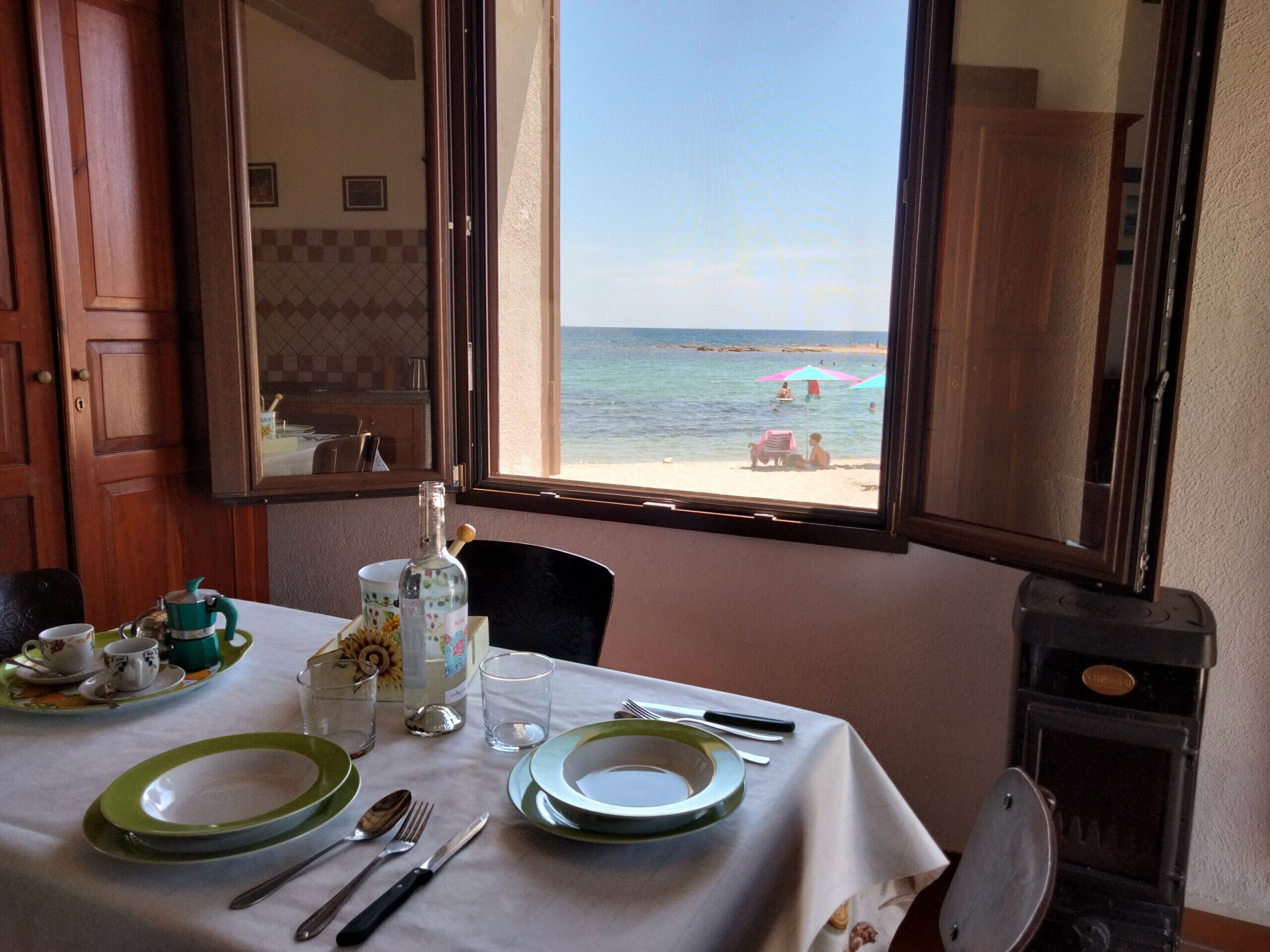 Ein Esstisch ist gedeckt mit Tellern, Besteck und einer Flasche, mit Blick auf den Strand durch ein offenes Fenster.