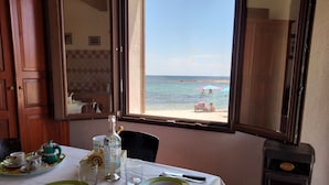 Ein Esstisch ist gedeckt mit Tellern, Besteck und einer Flasche, mit Blick auf den Strand durch ein offenes Fenster.