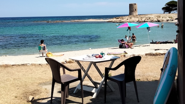 Ein Strand mit Tisch und Stühlen, einem Surfbrett und Menschen im Wasser.