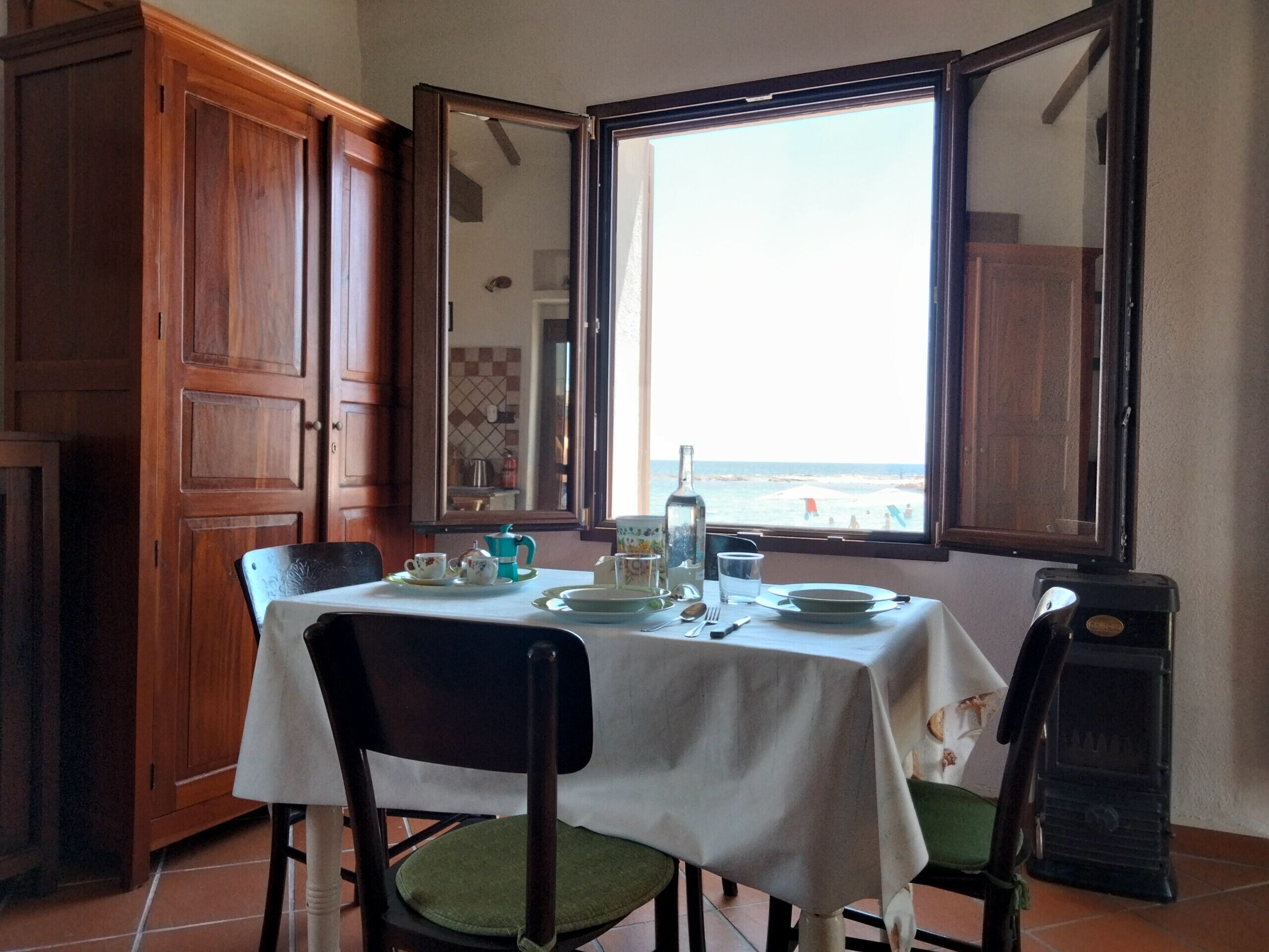 A dining area with a table set for two, a view of the beach through an open window, and a wooden cabinet to the left.