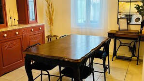 Uma área de refeições com mesa e cadeiras de madeira, luminária pendente, janela com cortinas e vista para uma lavanderia.