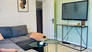 Uma sala de estar moderna com um sofá cinzento, uma mesa redonda pequena, uma estante de livros, uma TV sobre um suporte e um candelabro.