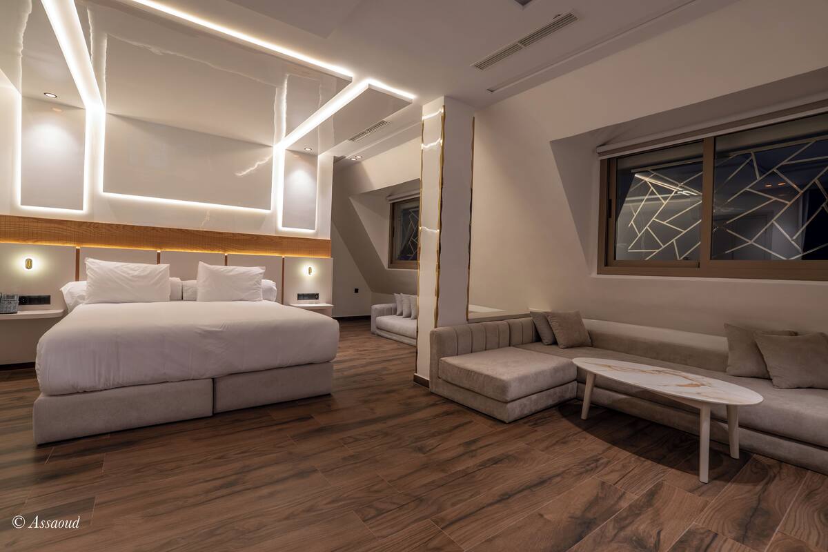 A modern hotel room with a large bed, a sofa, a coffee table, and a view of the cityscape.