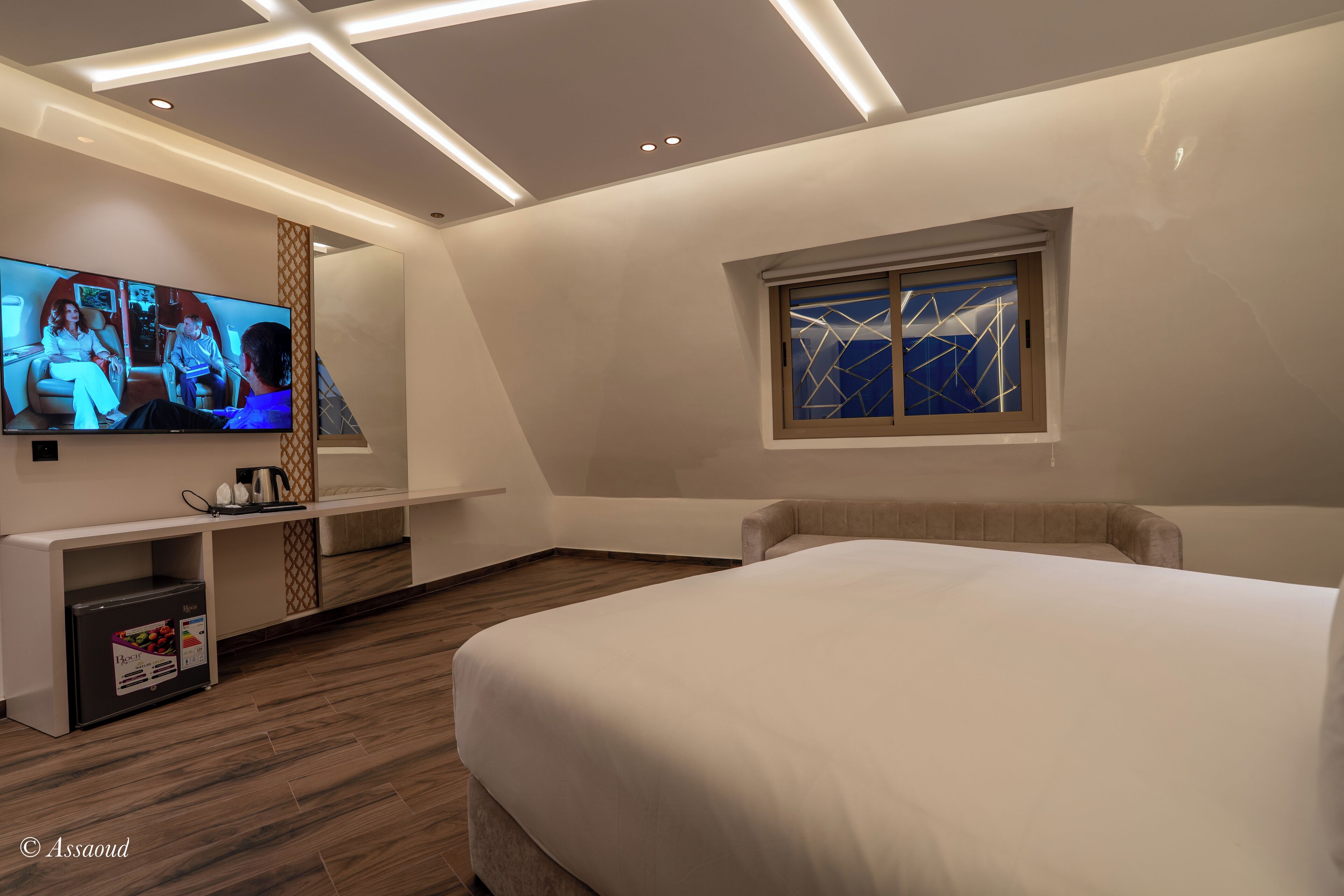 A modern hotel room with a large bed, a flat-screen TV, a small kitchenette, and a window with a geometric view.