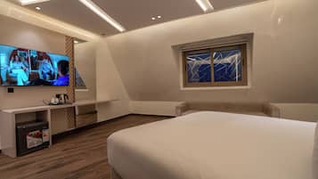 A modern hotel room with a large bed, a flat-screen TV, a small kitchenette, and a window with a geometric view.