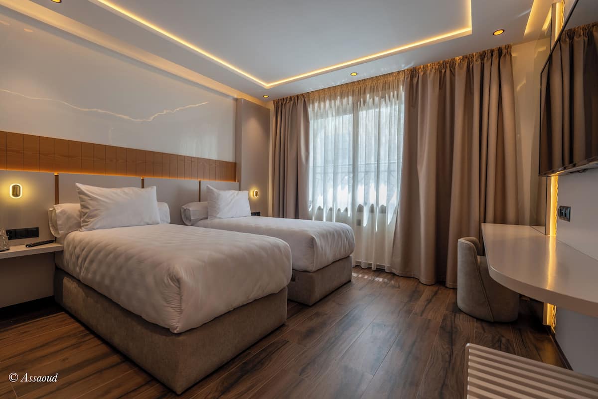 A modern hotel room with two beds, a flat-screen TV, a desk, and a large window with curtains.
