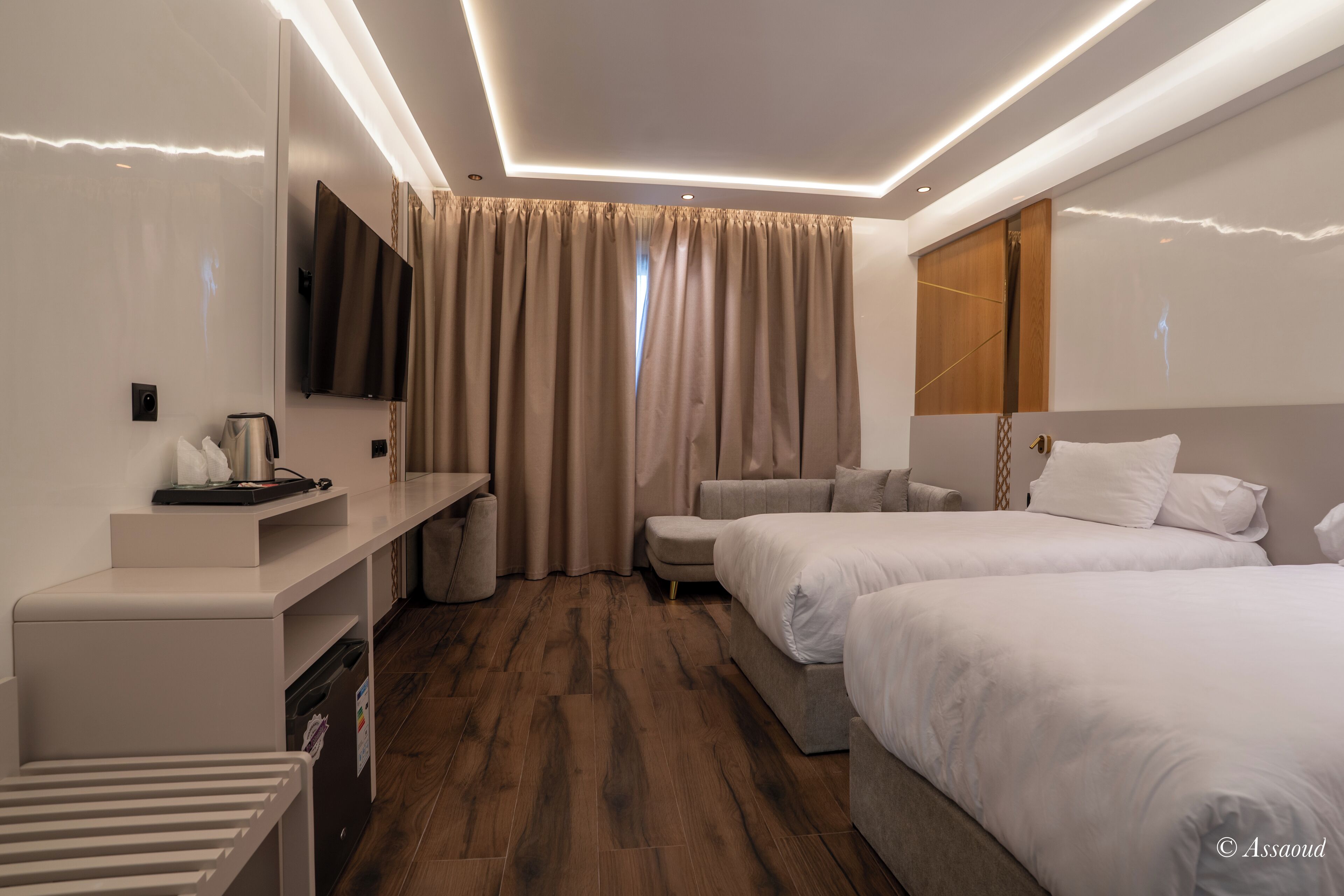 A hotel room with a flat-screen TV, a desk with a coffee maker, two beds, a sofa, and a large window with curtains.