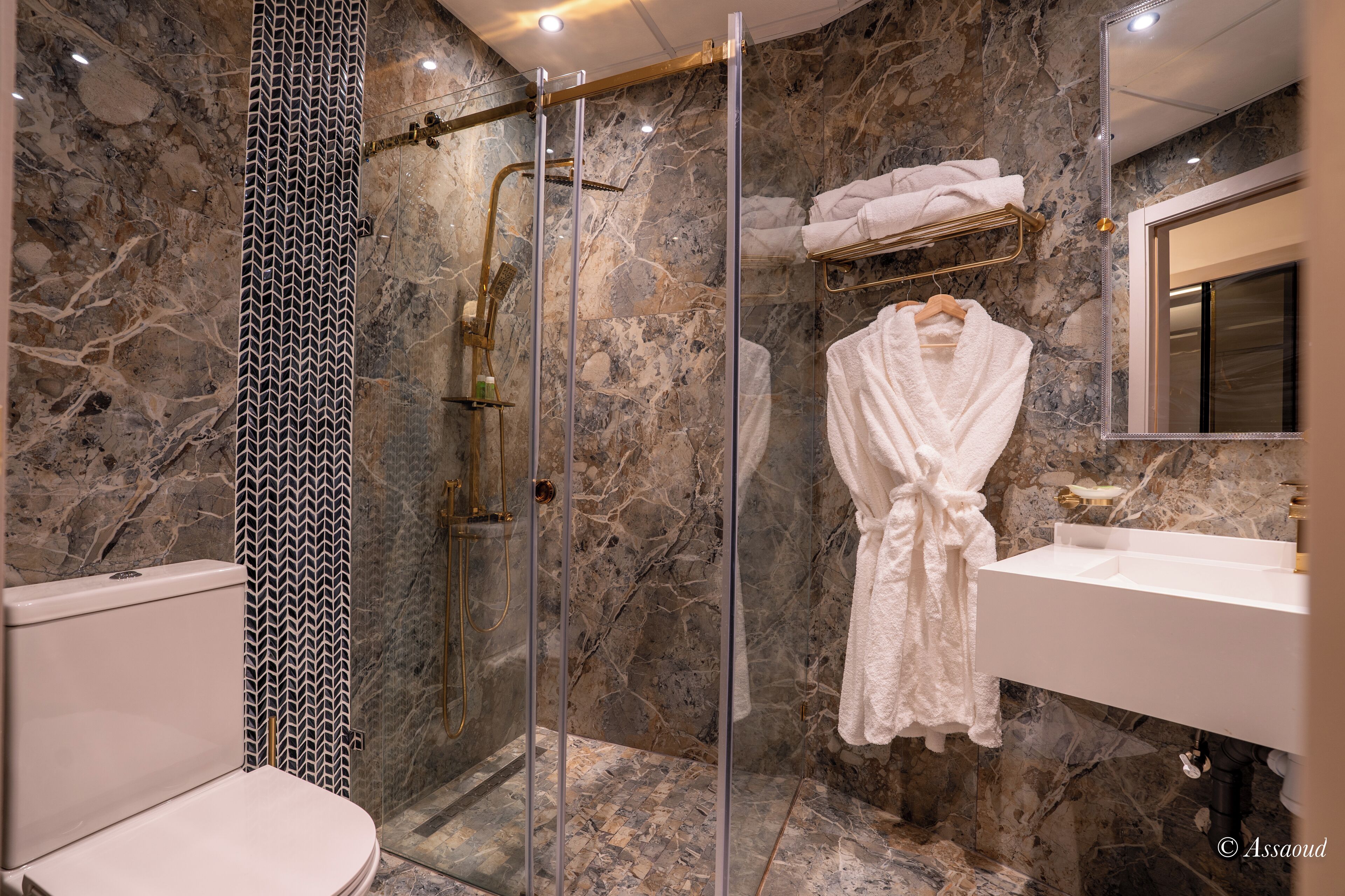 Un baño moderno con ducha de mármol, ducha a pie con mampara de vidrio, lavamanos blanco y una bata colgada en un perchero.