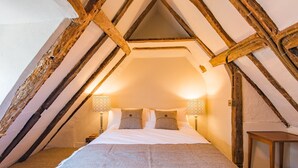 Um quarto de sótão aconchegante, com teto inclinado, uma cama com cobertor cinza, duas luminárias de cabeceira e uma pequena mesa de madeira.