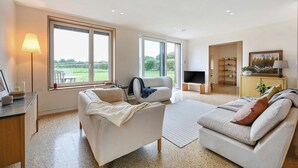 Un salon spacieux avec deux canapés, une table basse et un coin télévision. De grandes fenêtres offrent une vue sur l’extérieur.
