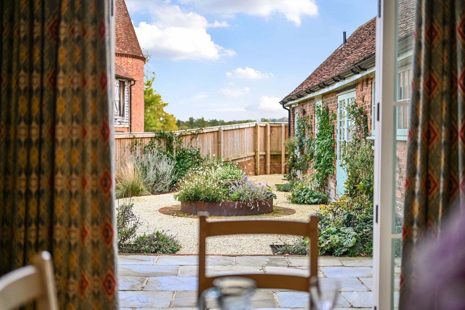 A view from inside a room looking out to a garden with a bench, a wooden fence, and a brick building.
