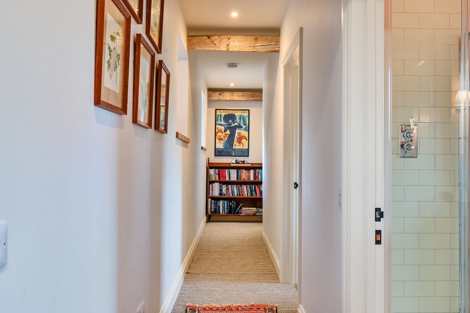 Um corredor com quadros nas paredes à esquerda, uma estante com livros e uma porta que dá para o banheiro.