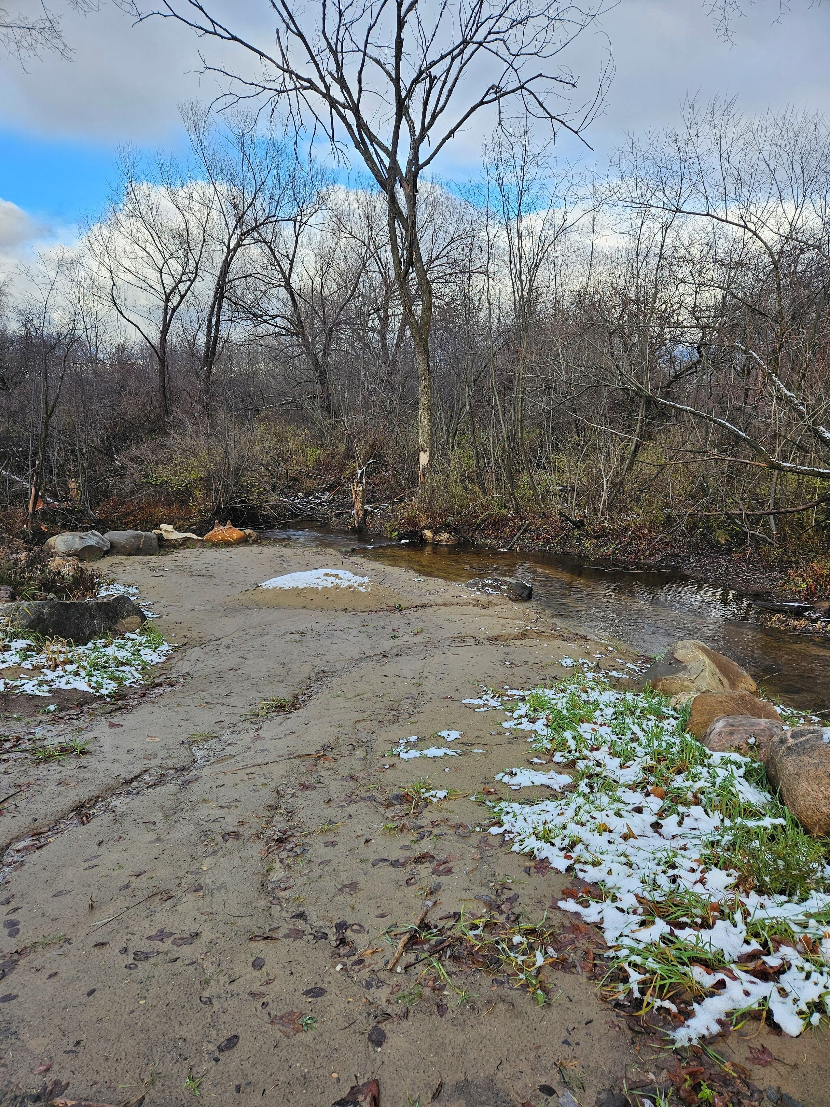 A natural landscape with a stream, bare trees, and patches of snow on the ground.