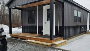 A single-story metal building with a covered porch, a front door, and a small window. - Wonderful 1-bedroom cabin with AC in charming Potsdam on a brook. (Potsdam)