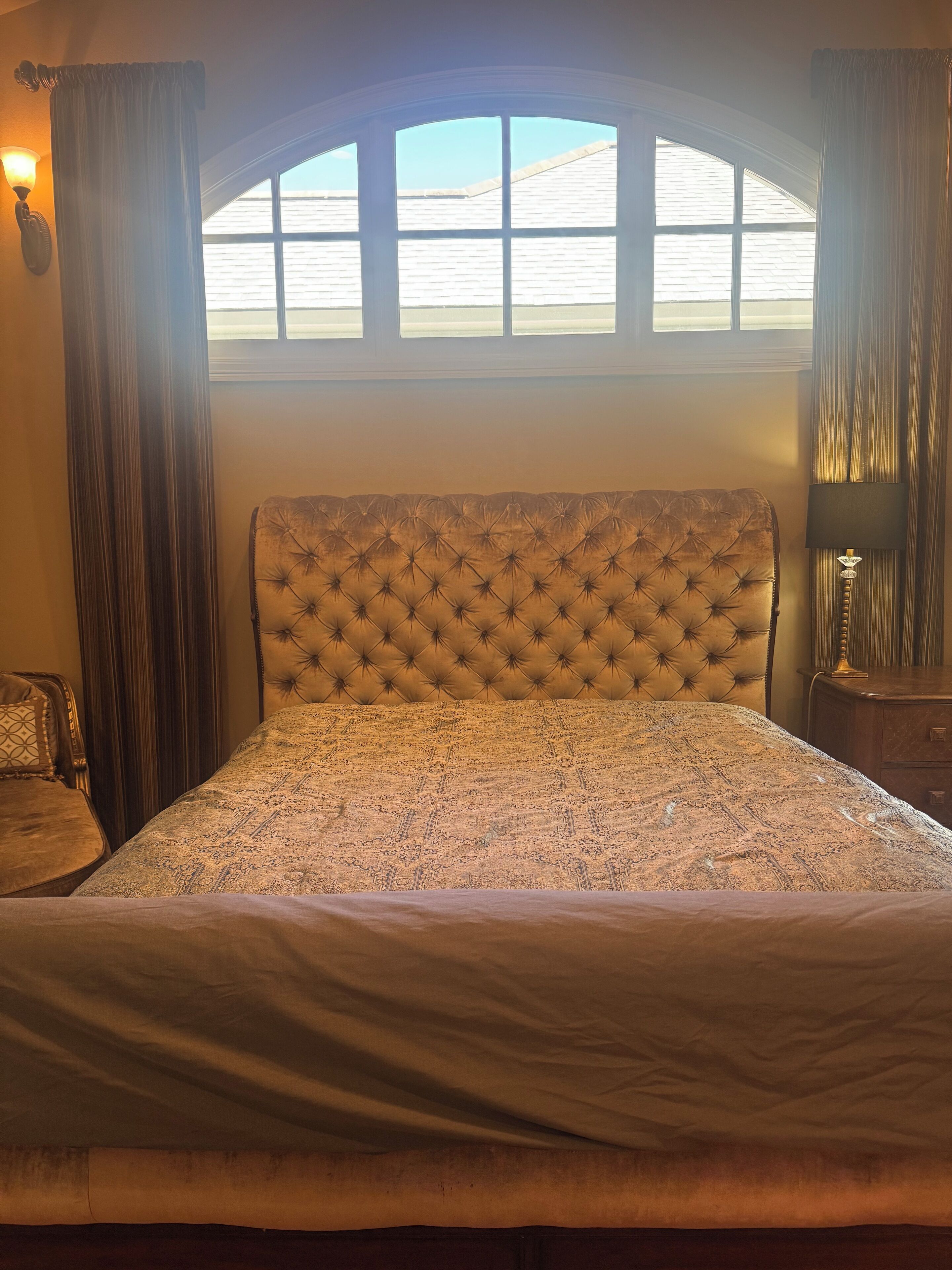 A bedroom with a tufted headboard, a patterned bedspread, and a window with curtains.