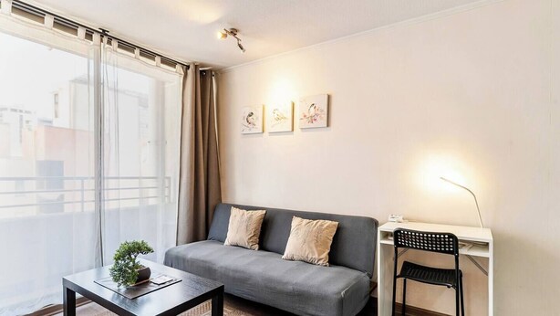 A modern living room with a grey sofa, a small table with a plant, a desk with a chair, and a balcony with a view of buildings. - Apartment in Metro U. de Chile with Balcony, Gym & Swimming Pool, Historic Center Santiago (Santiago)
