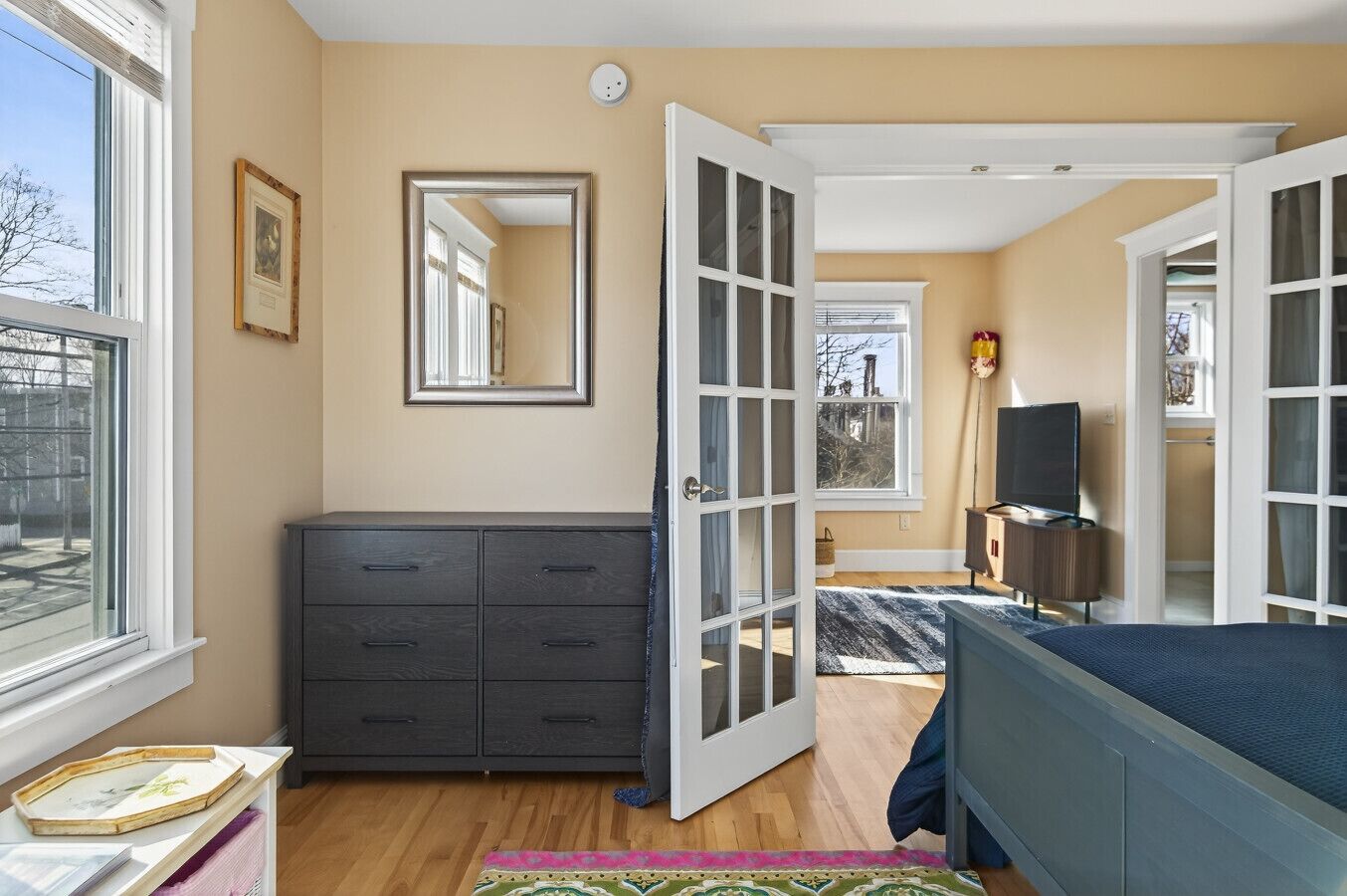 A bedroom with a dark dresser, a bed with a blue cover, a TV, and a large window.
