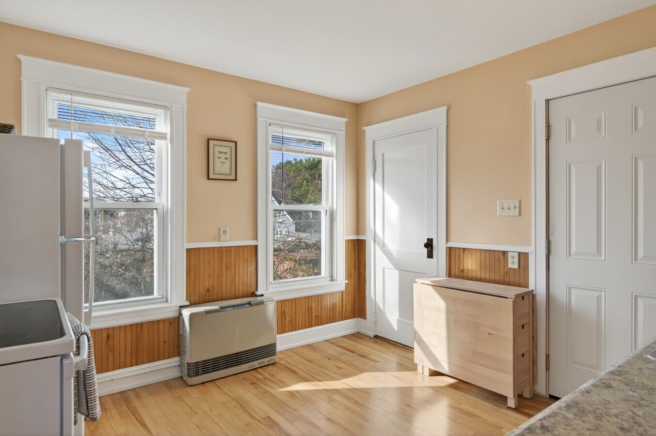 Una cocina con pisos de madera, gabinetes blancos, horno empotrado y ventana con vista a árboles.