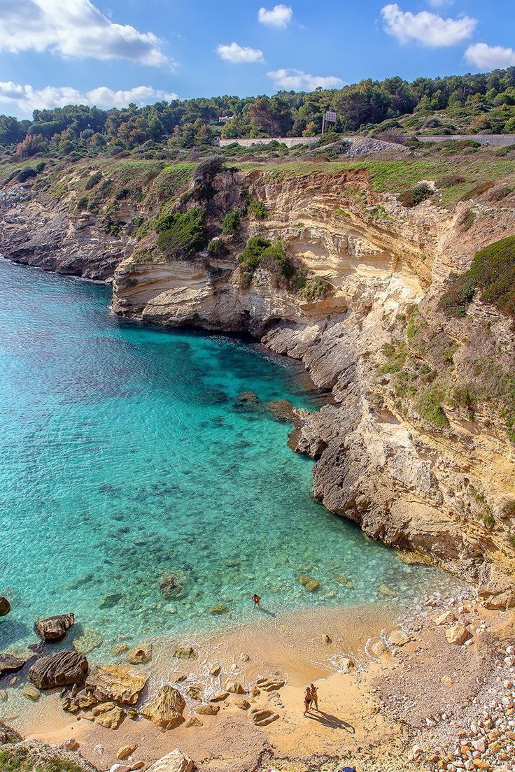 A coastal landscape with clear turquoise waters, rocky cliffs, and a sandy beach.