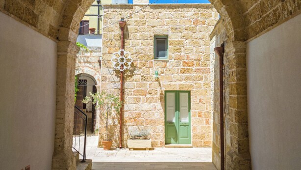 Un corredor de piedra con arco, una puerta verde y un ventanal pequeño, que conduce a un edificio con tejado de azulejos.