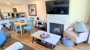 A modern living room with a fireplace, a dining area with a round table and chairs, and a kitchen in the background.