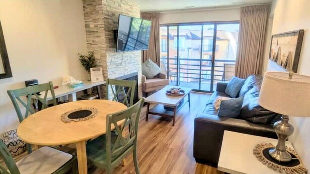 A modern living room with a stone fireplace, a dining area with a round table and chairs, a sofa, and a TV mounted on the wall.