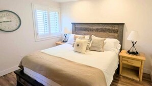 A bedroom with a bed, a nightstand, a lamp, a clock, and a window with blinds.