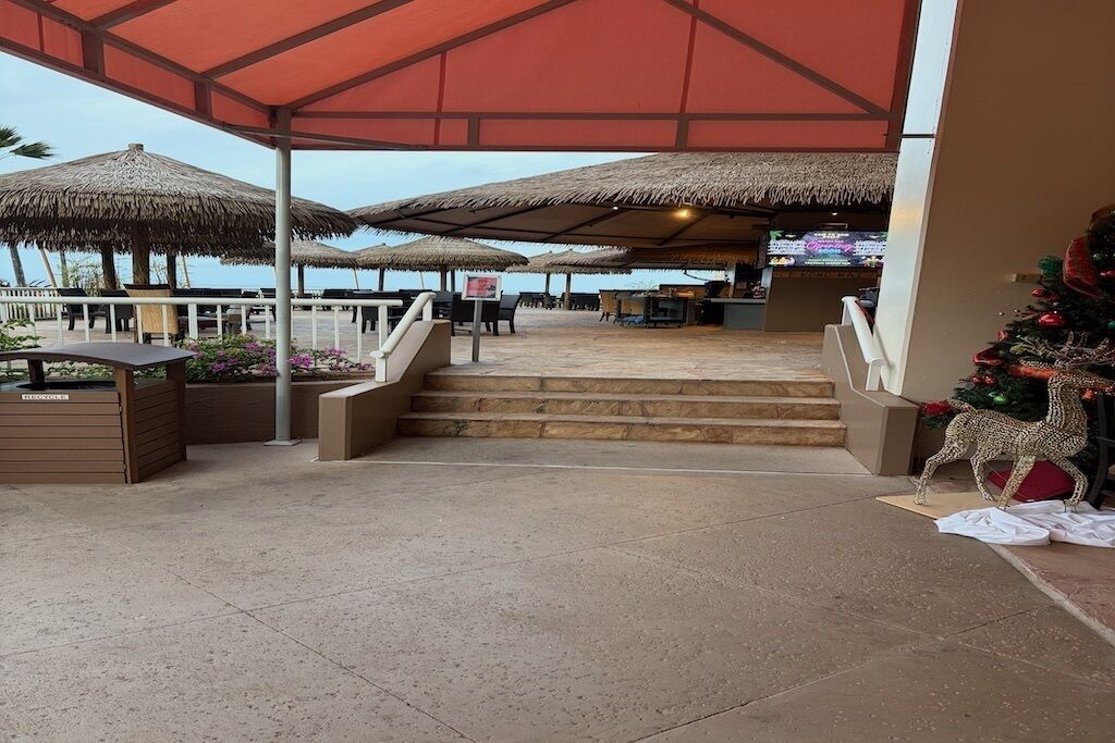 Un espacio al aire libre techado con sombrillas de paja, asientos y una escalera que lleva a una zona de bar.