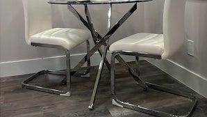 Un comedor con una mesa de vidrio, dos sillas blancas, una planta en maceta y un cuadro en la pared.