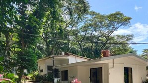 Un bâtiment d’un seul étage, doté d’un toit plat, entouré d’un jardin où poussent différentes plantes et arbres.