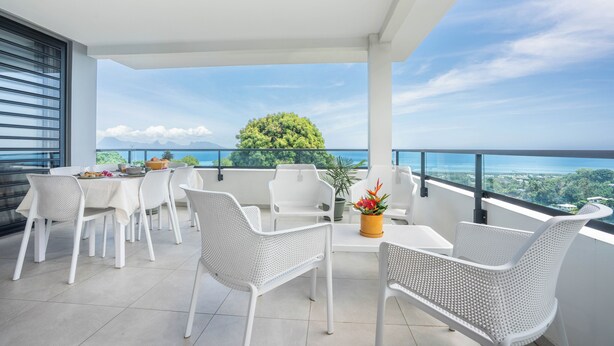 Un balcone con mobili in vimini bianco, un tavolo con frutta e vista sull'oceano e sul verde.