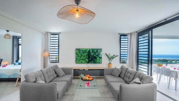A modern living room with a large flat-screen TV, grey sofas, a glass coffee table, and a view of the ocean through large windows.
