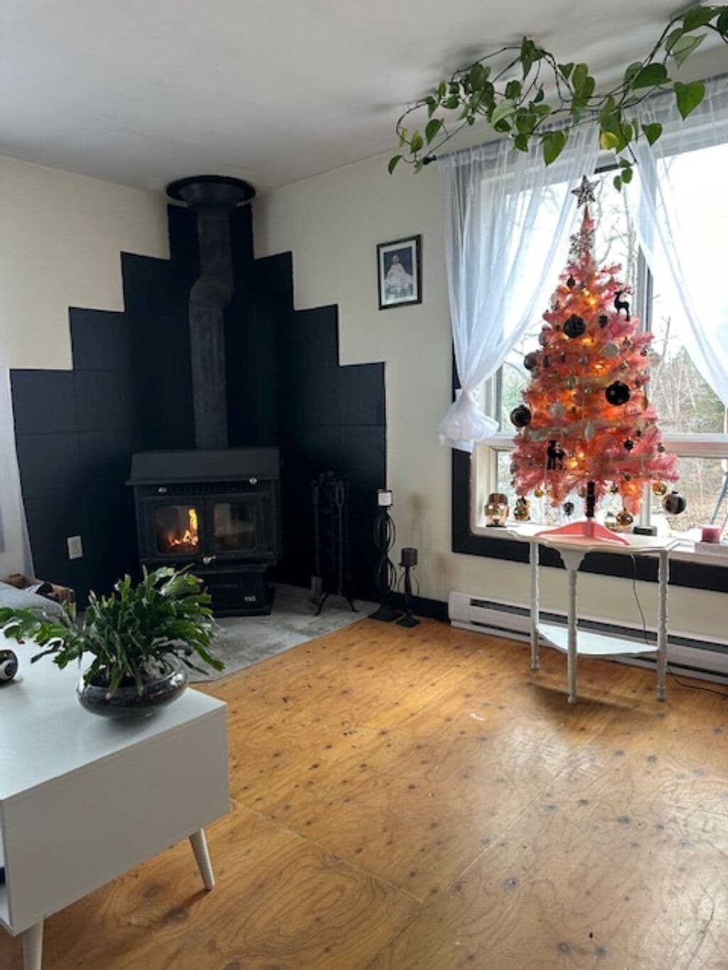 A cozy living room with a wooden floor, a fireplace, a Christmas tree, and a window with curtains.