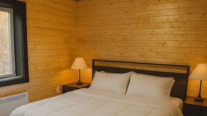 Un dormitorio con piso de madera, una cama con ropa de cama blanca, dos mesitas de noche con lámparas y una ventana con vistas.