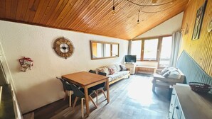 Une pièce chaleureuse avec un plafond en bois, une table à manger avec des chaises, un canapé, un téléviseur et une fenêtre avec des rideaux.