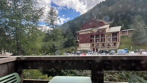 Une terrasse avec des chaises et une table en métal vert, donnant sur un parking et un immeuble doté de nombreux balcons, le tout sur fond d’arbres et d’un ciel nuageux.