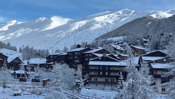 Une station de montagne enneigée, avec plusieurs bâtiments en bois, dont un grand chalet doté de balcons, et un télésiège visible au loin.