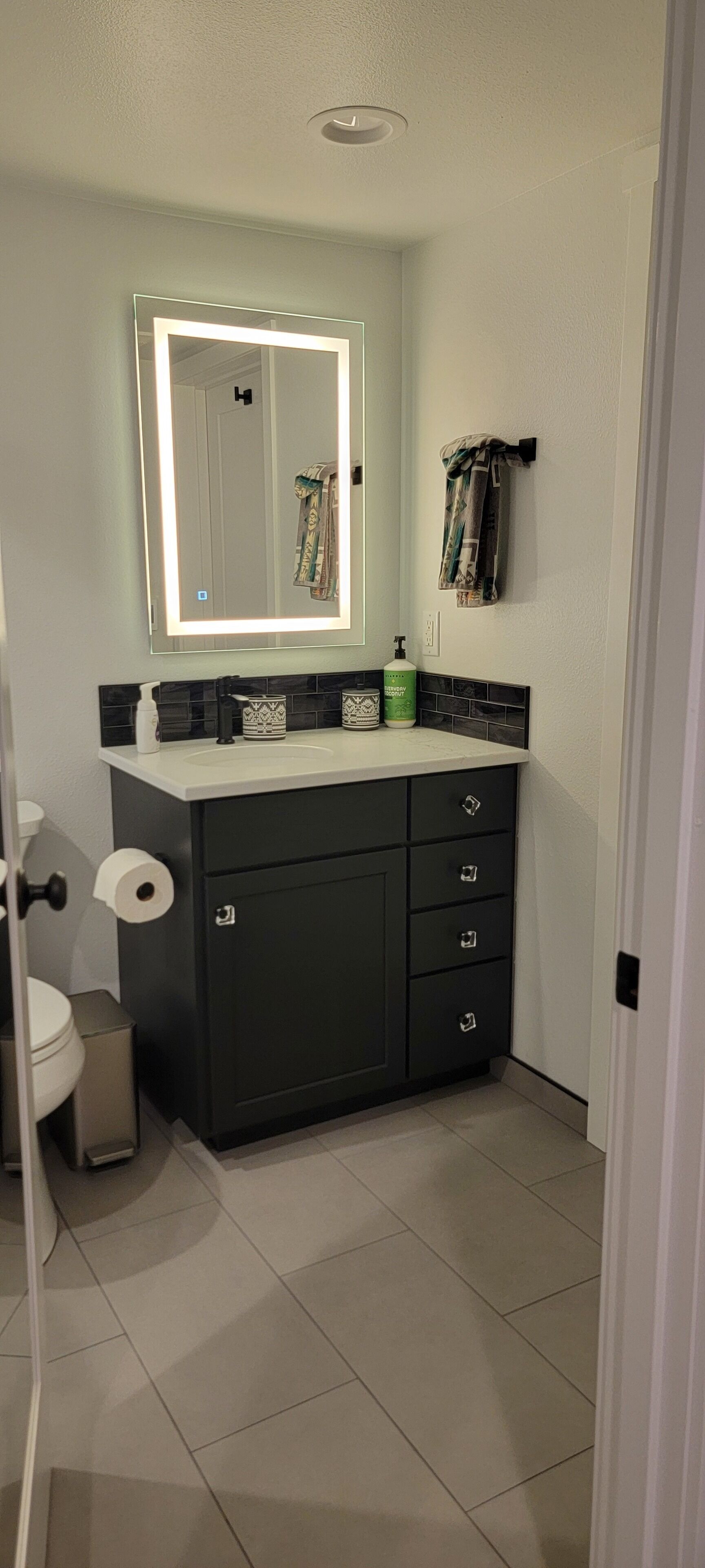 Un baño con un mueble de tocador oscuro, un espejo con luz, un inodoro y un perchero para toallas.
