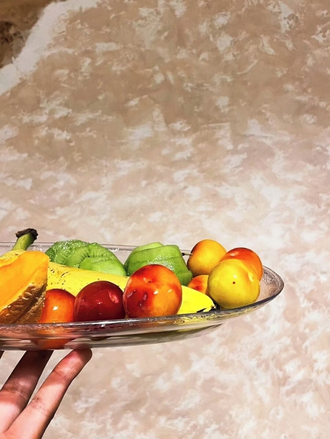 A hand holding a glass plate with various fruits including a slice of mango, kiwi, green apple, and peaches.